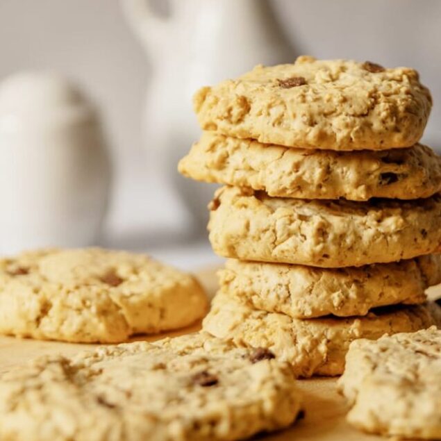 A stack of oatmeal chocolate chip cookies on a wooden board, with additional cookies nearby.