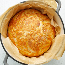 A golden-brown loaf of Cheese Bread in a Dutch oven, lined with parchment paper.