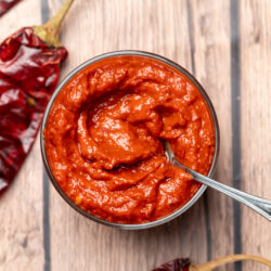 Homemade harissa paste in a glass bowl with a spoon, surrounded by dried red chiles.