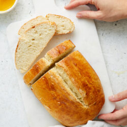 Partially sliced Italian bread on a white cutting board, held by a person's hands.