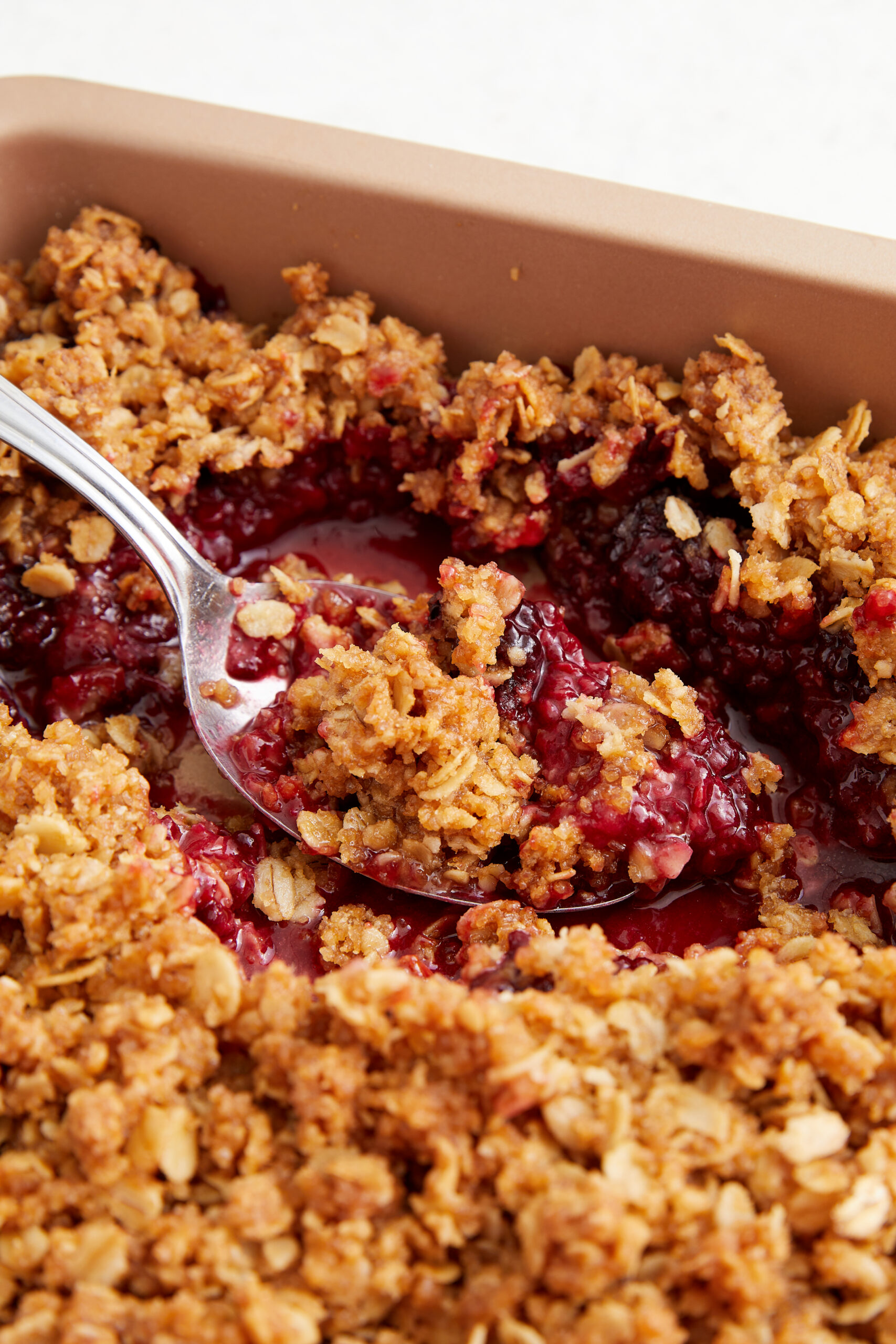 A spoon scooping a warm blackberry crisp from a brown baking dish.