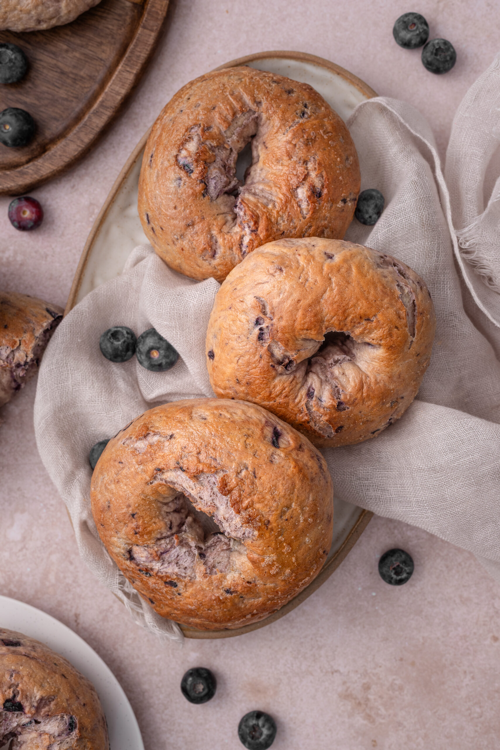 Three freshly baked blueberry bagels on a plate with scattered fresh blueberries.