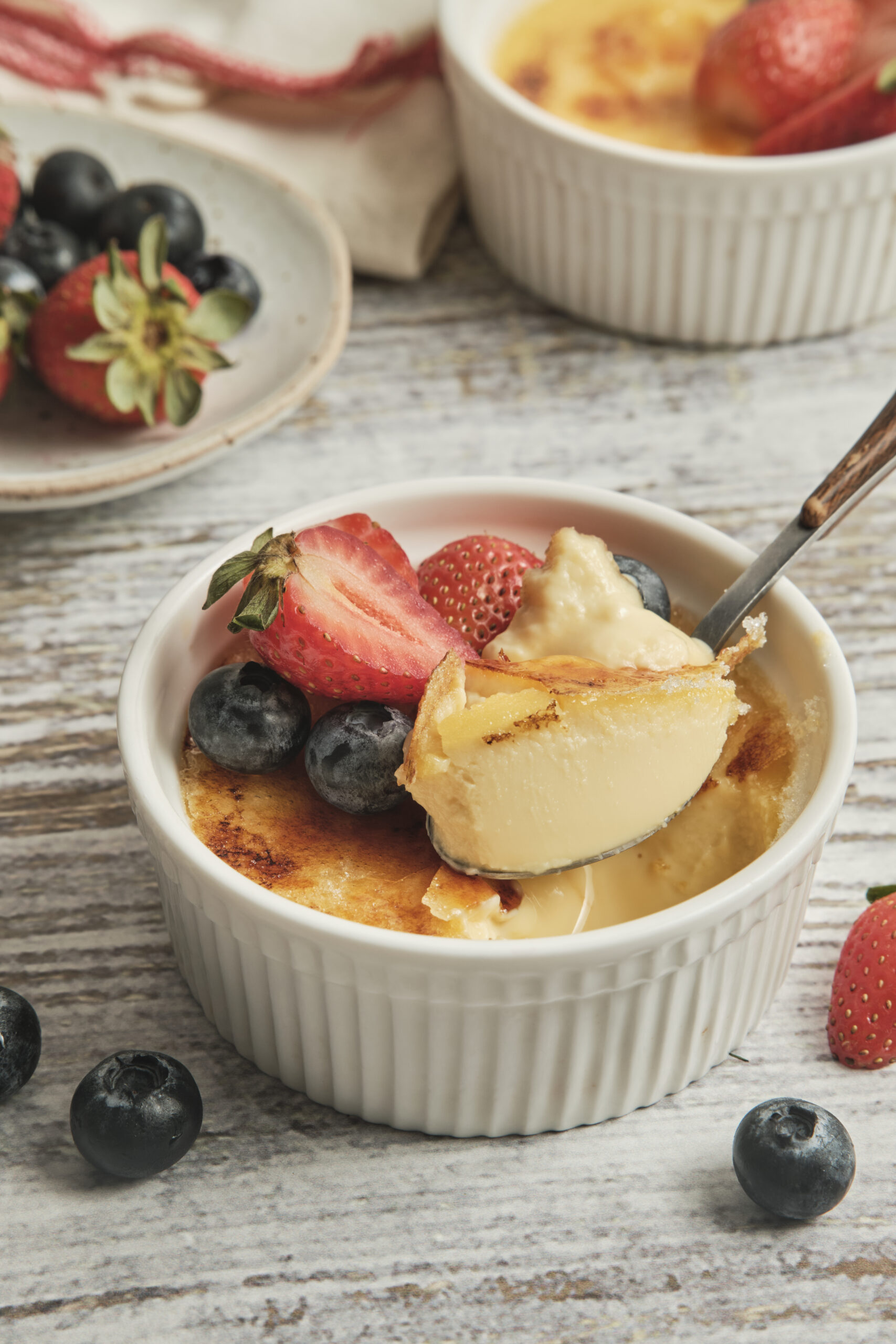 Cr&egrave;me br&ucirc;l&eacute;e in a white ramekin, garnished with fresh strawberries and blueberries, with a spoon.
