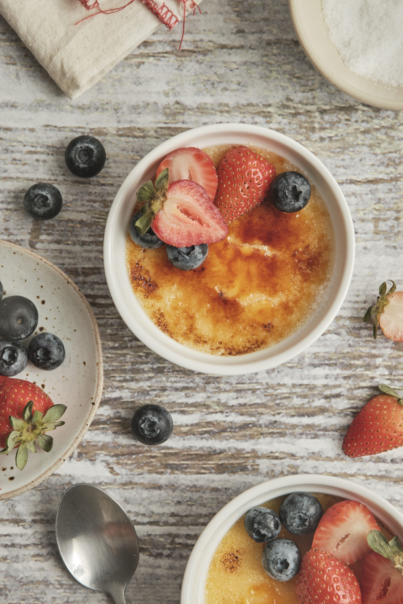 Cr&egrave;me br&ucirc;l&eacute;e in white ramekins, garnished with fresh strawberries and blueberries.