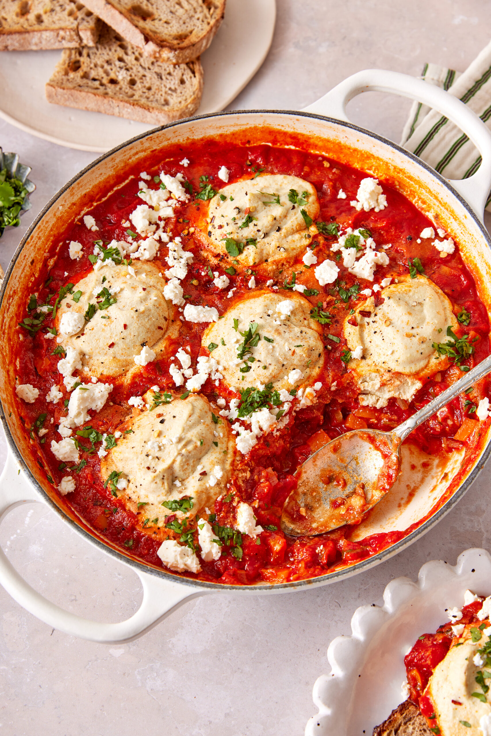 Vegan shakshuka in a white skillet, topped with vegan feta, cilantro, and crusty bread.