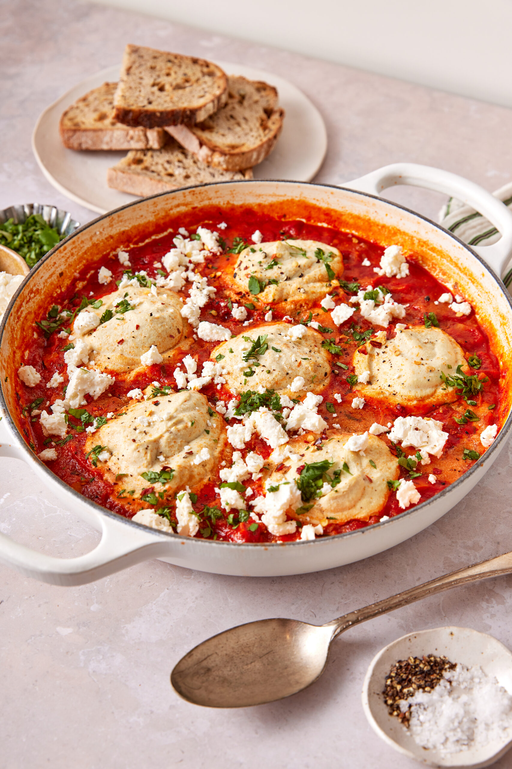 Vegan shakshuka in a white skillet, topped with tofu, vegan feta, and cilantro, served with bread.