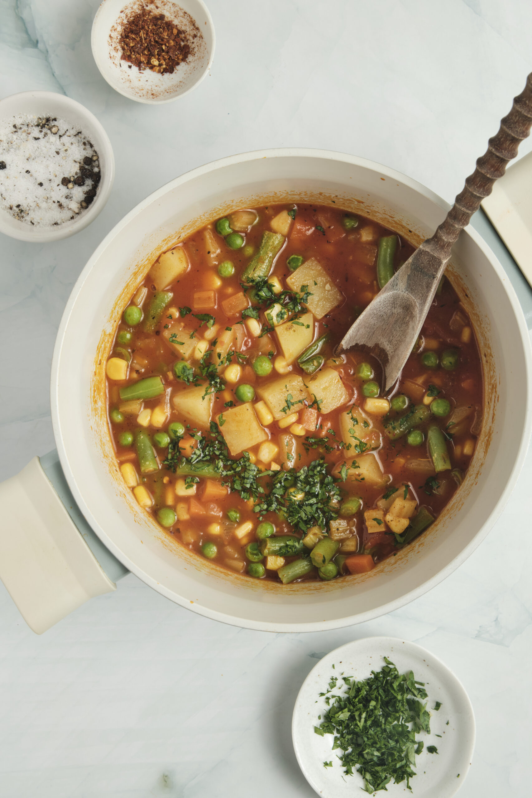 Vegetable soup in a white pot with mixed vegetables, garnished with parsley.