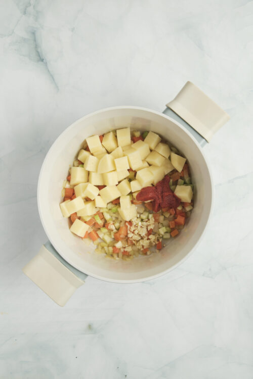 Adding diced potatoes, minced garlic, and tomato paste to the pot for vegetable soup.